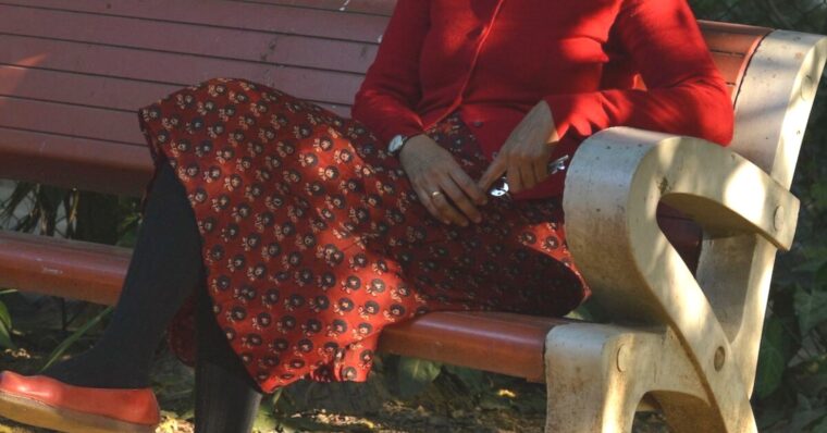 Close up of a woman sitting on a bench wearing a red Garibaldi Skirt, red button-up cardigan and red flats